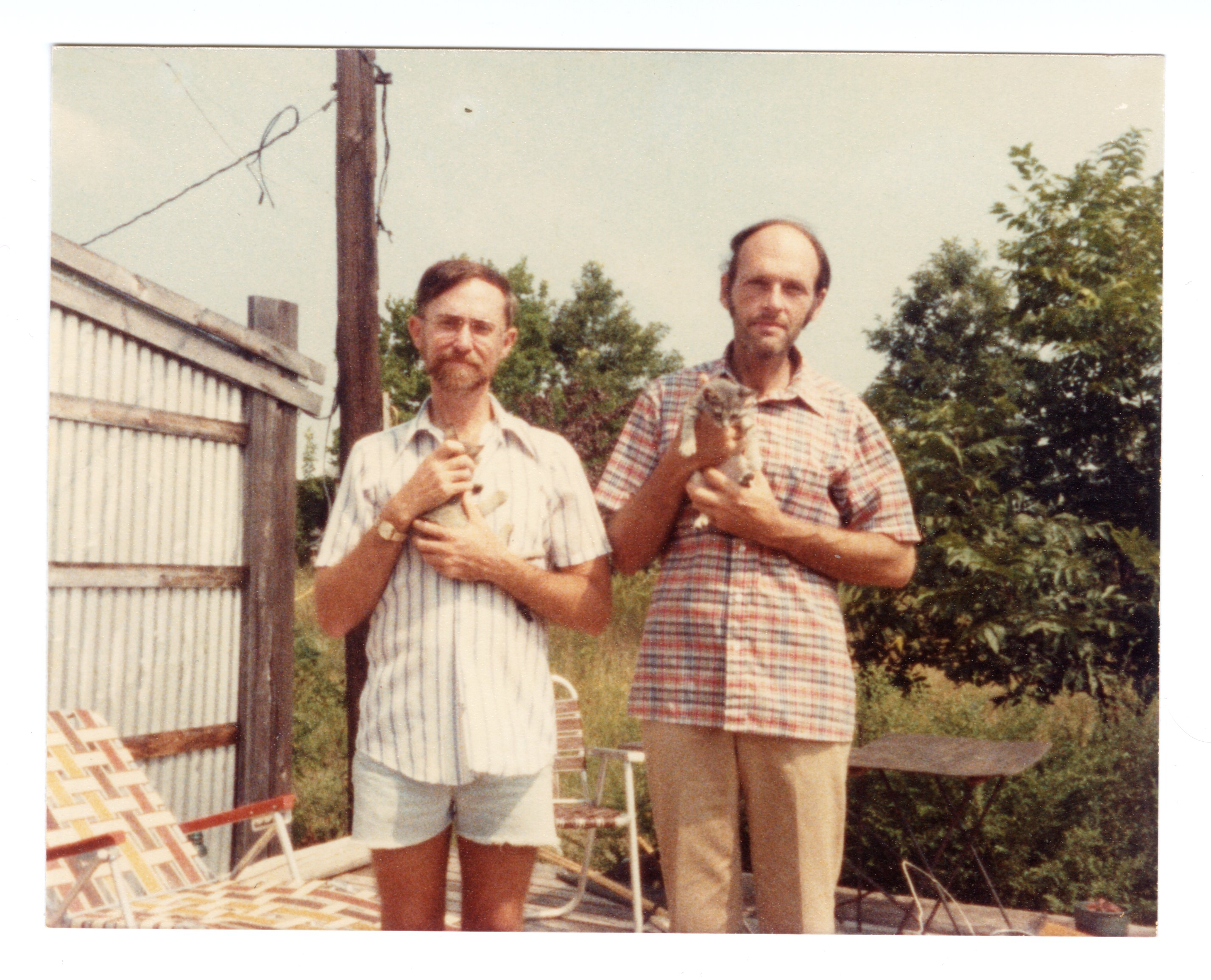 Two men stand in front of a fence and are holding small kittens.