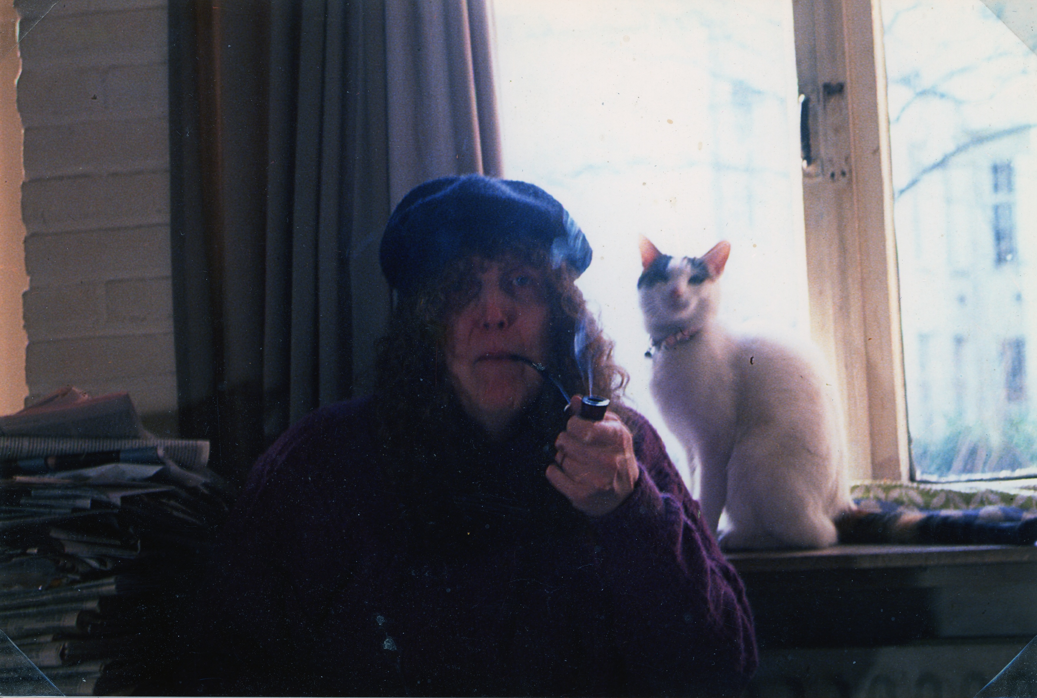 A woman (Hennix) smokes a pipe and sits in front of a cat and a curtain.