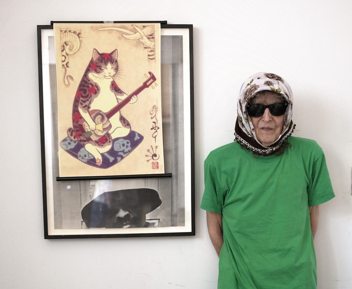 Catherine Christer Hennix wears a green t shirt, a head scarf, and glasses. She poses in front of a poster of a cat playing an instrument.