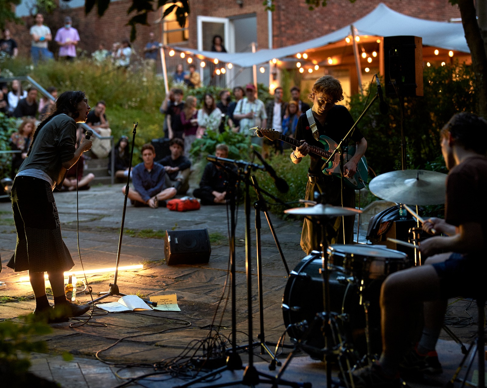 a singer, guitarist, and drummer perform in a dim garden under a spotlight.