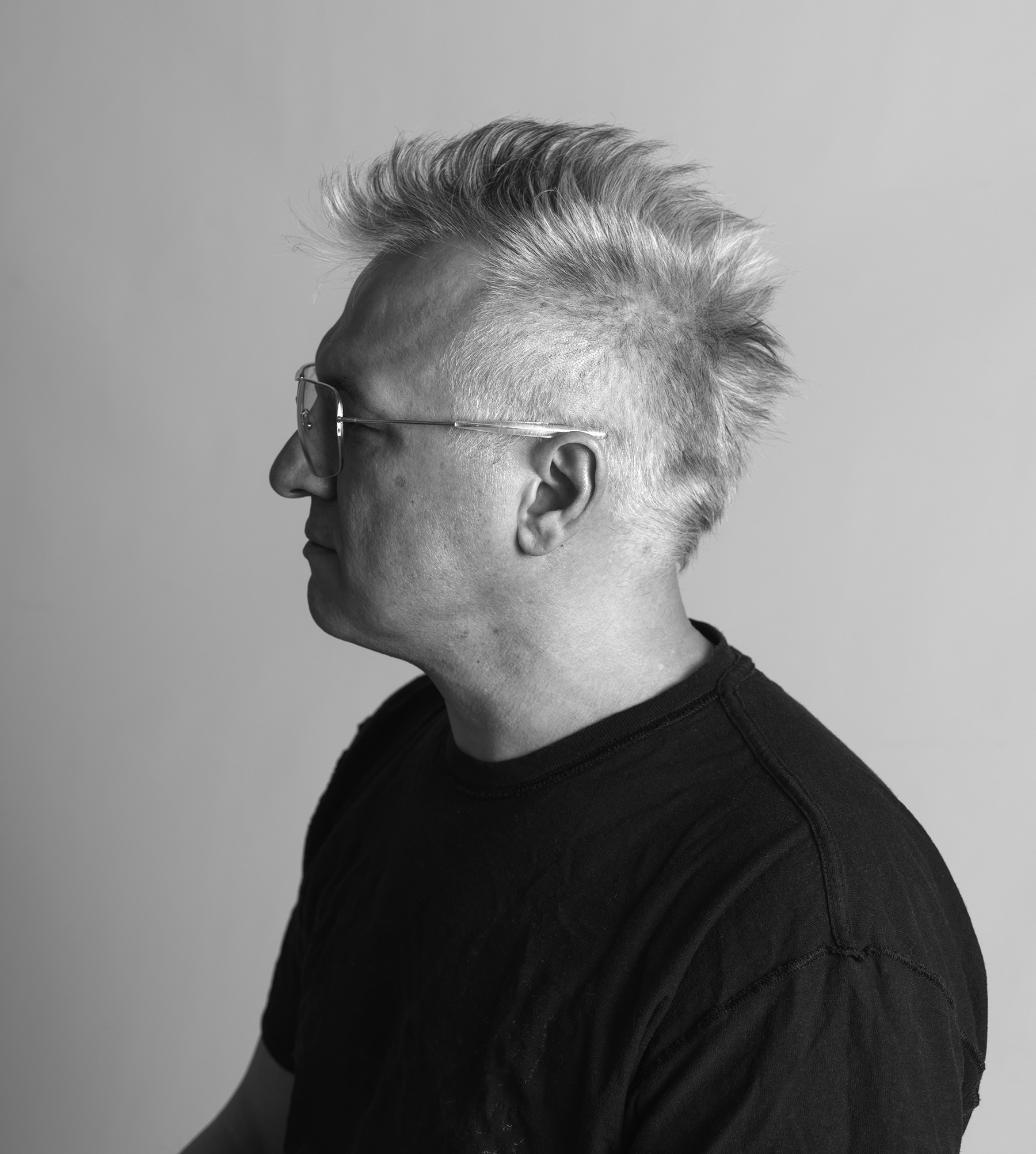 A man with a silver mohawk looks to the left, wears a black shirt and glasses.
