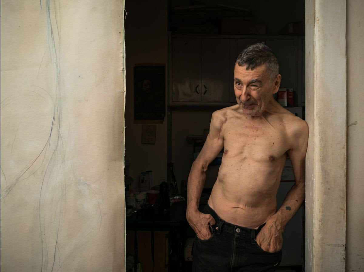 Loren Connors standing shirtless in a doorway next to a painting