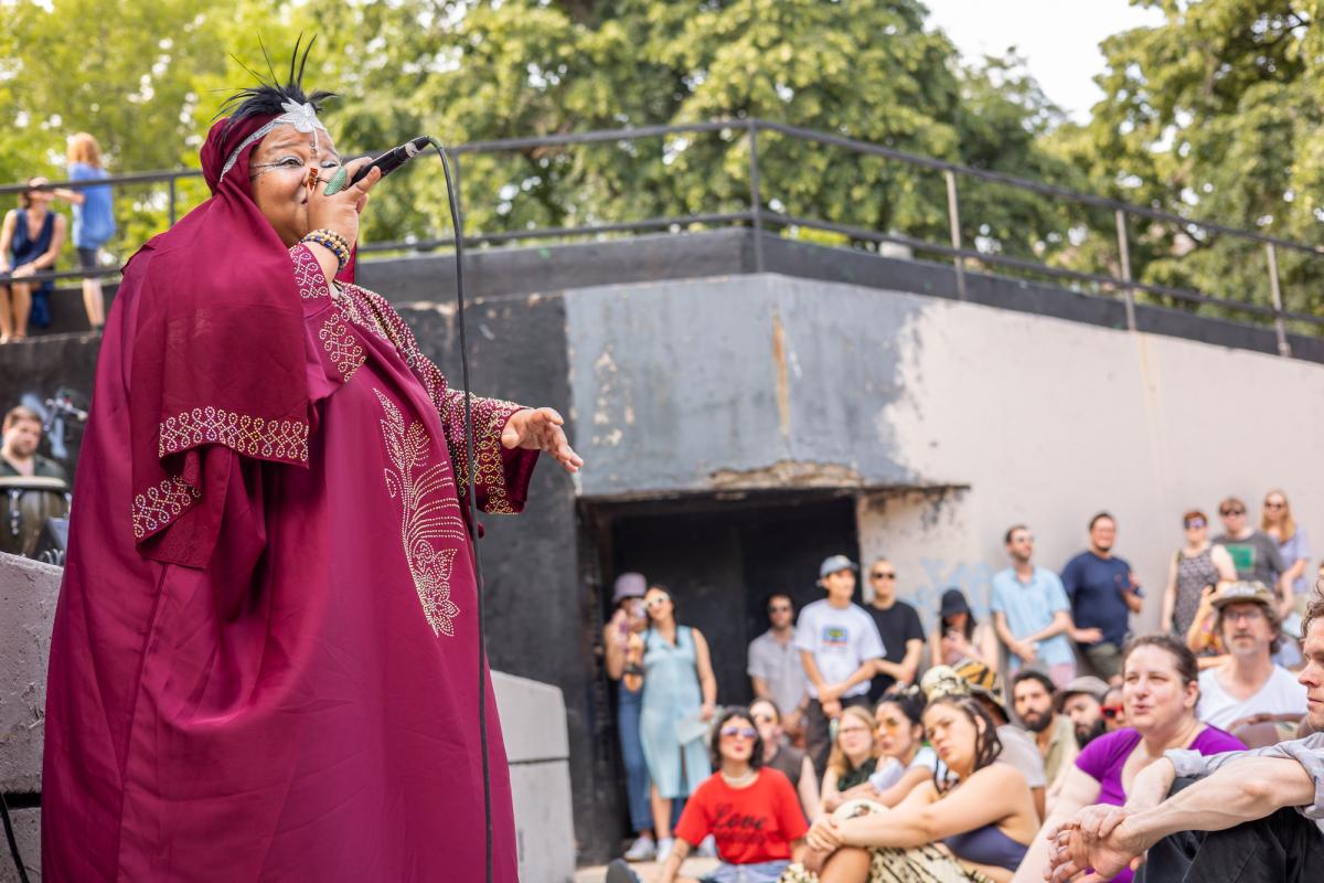A woman in a burgundy robe (Angel Bat Dawid) sings in front of an outdoor audience.