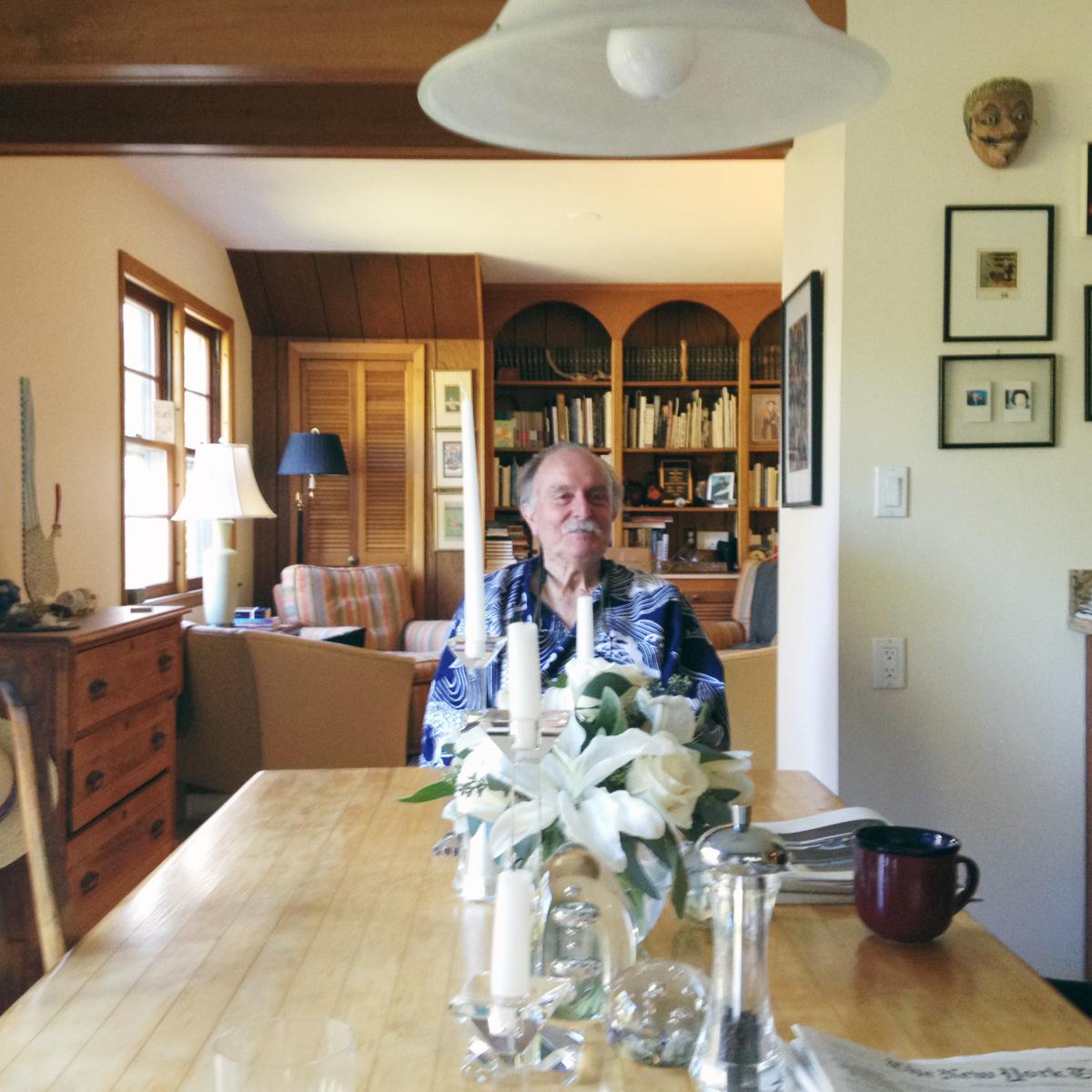 Alvin Lucier sits at a table with lilies and a coffee mug in front of him.