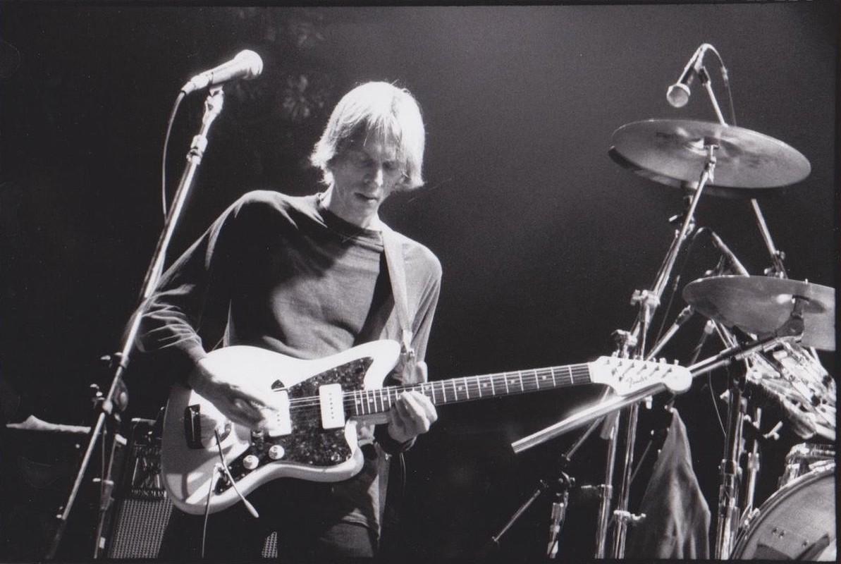 Tom Verlaine, a man with floppy blond hair, plays an electric guitar in front of a mic stand.